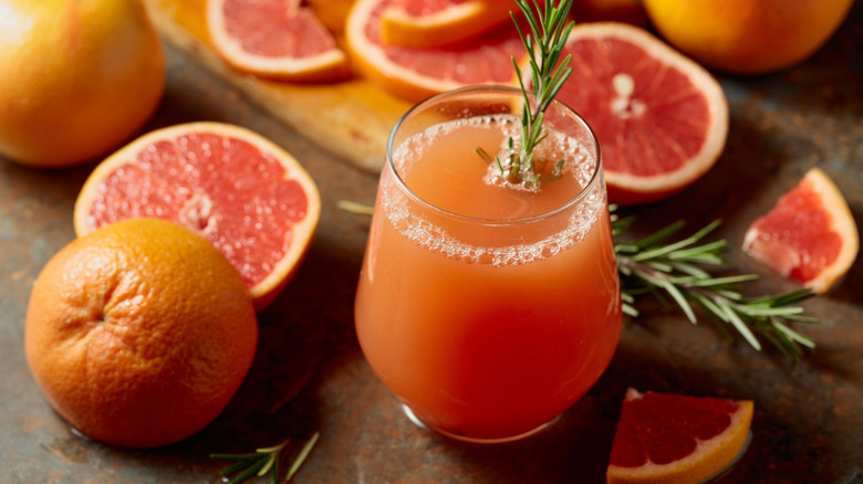 Een glas grapefruitsap met een rozemarijn tapje omgeven door hele en gesneden grapefruit