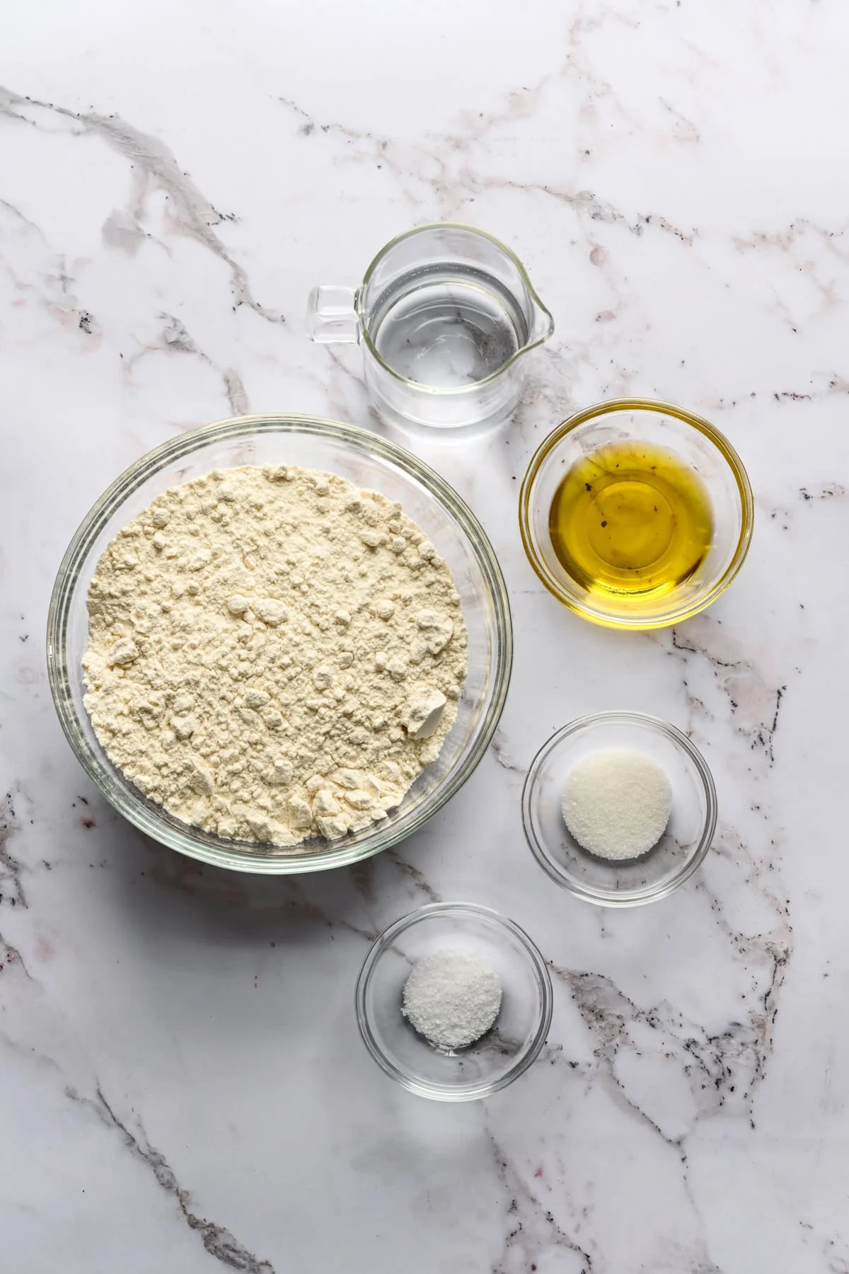 bovenaanzicht van bloem, water, olijfolie, zout en suiker in kommen.