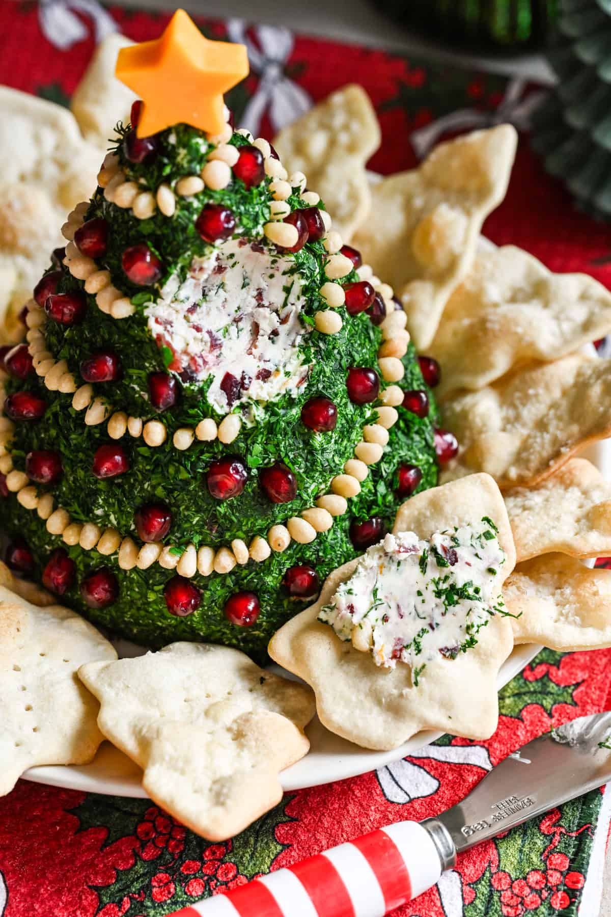 Kerstboomvormige kaasbal bedekt met kruiden en versierd met pijnboompittenslinger en granaatappelornamenten.
