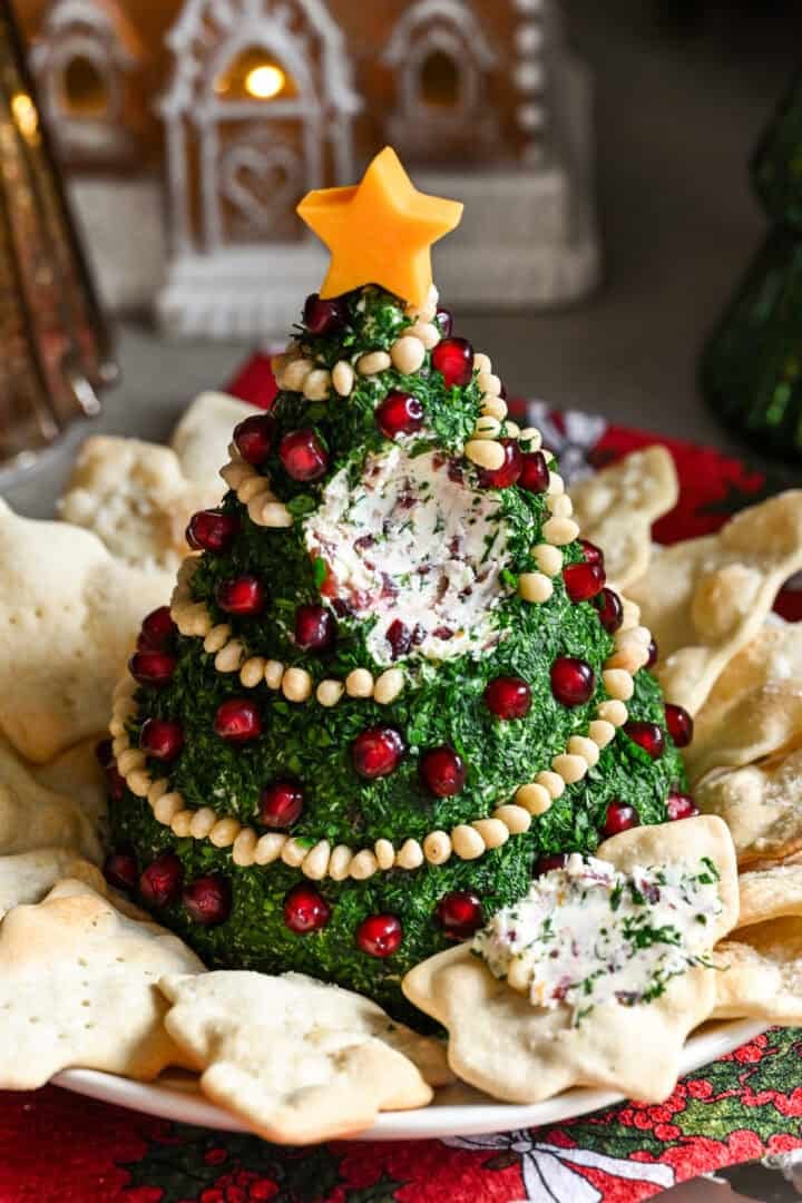 Kerstboomvormige kaasbal bedekt met kruiden en versierd met pijnboompittenslinger en granaatappelornamenten.