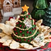 Kerstboomvormige kaasbal bedekt met kruiden en versierd met pijnboompittenslinger en granaatappelornamenten.