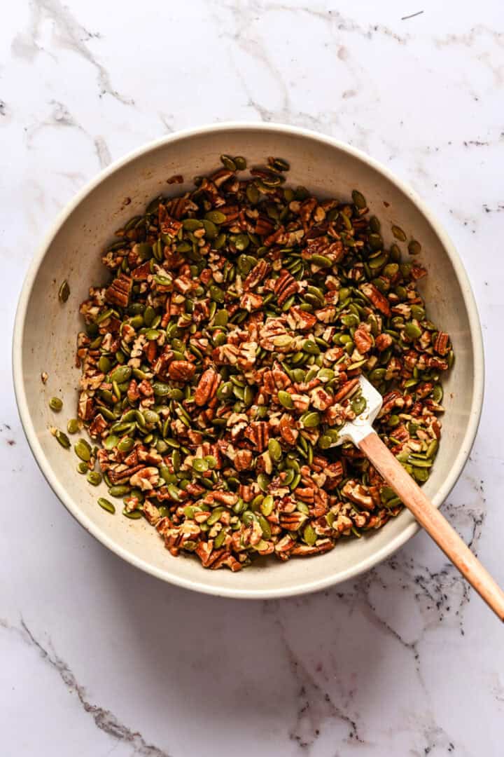 Pecannoten en pepitas samen gemengd in een kom met ahornsiroopmengsel.