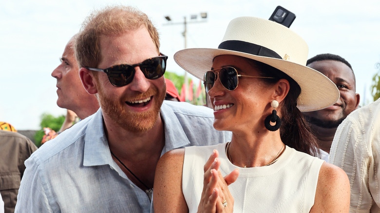 Prins Harry leunt over de schouder van Meghan Markle, beiden met een zonnebril.