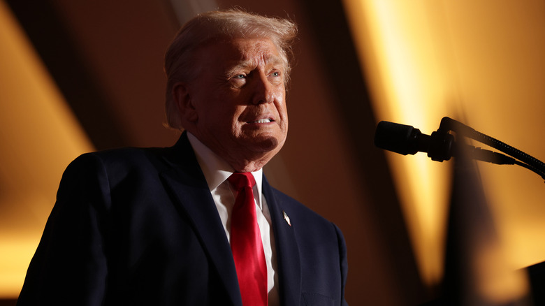 Donald Trump maakt een ineenkrimpende uitdrukking in een pak achter een podium, lage hoek.