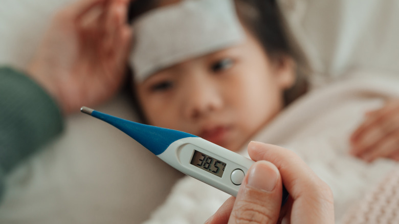 Een hand die een digitale thermometer vasthoudt terwijl een kind met koorts gaat liggen