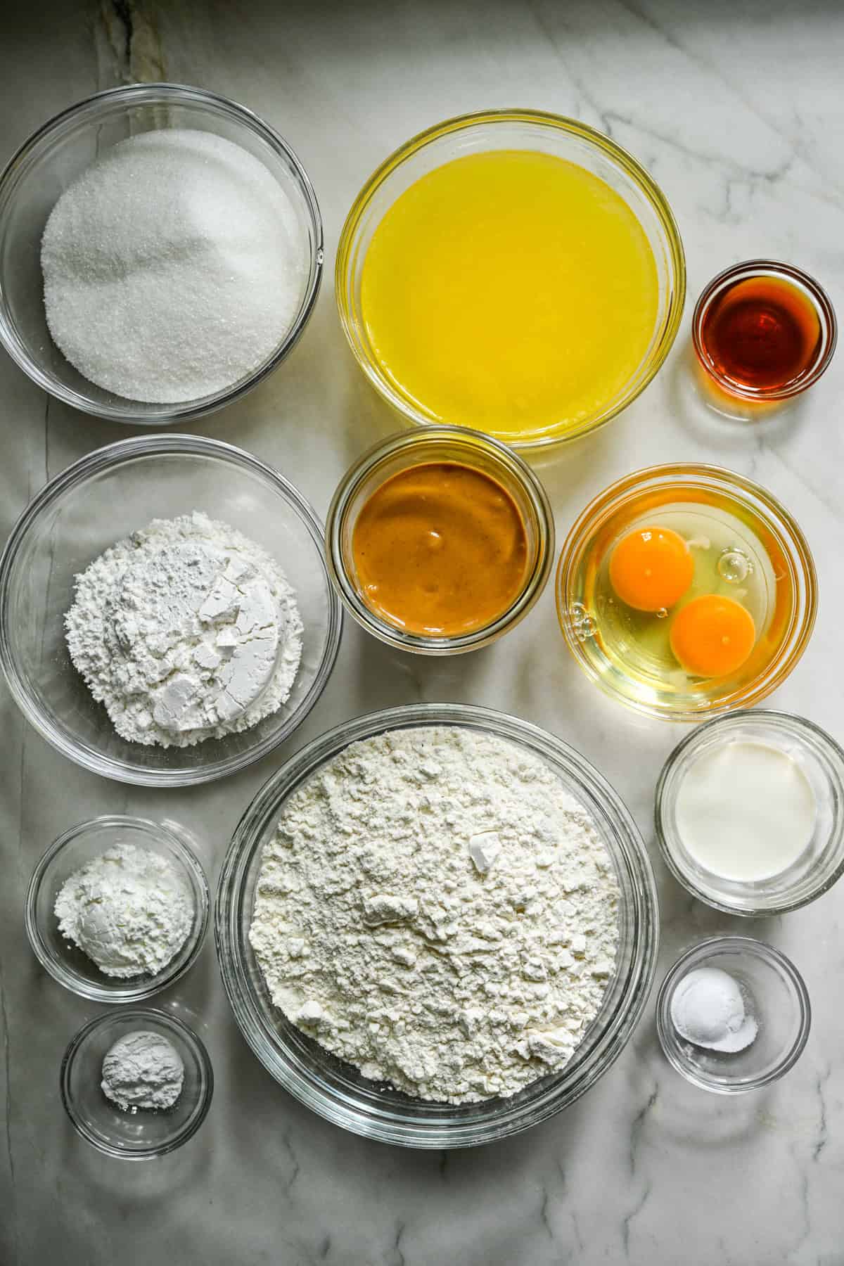 Bovenaanzicht van ingrediënten voor zelfgemaakte notenboters in aparte prep-kommen op wit aanrecht.
