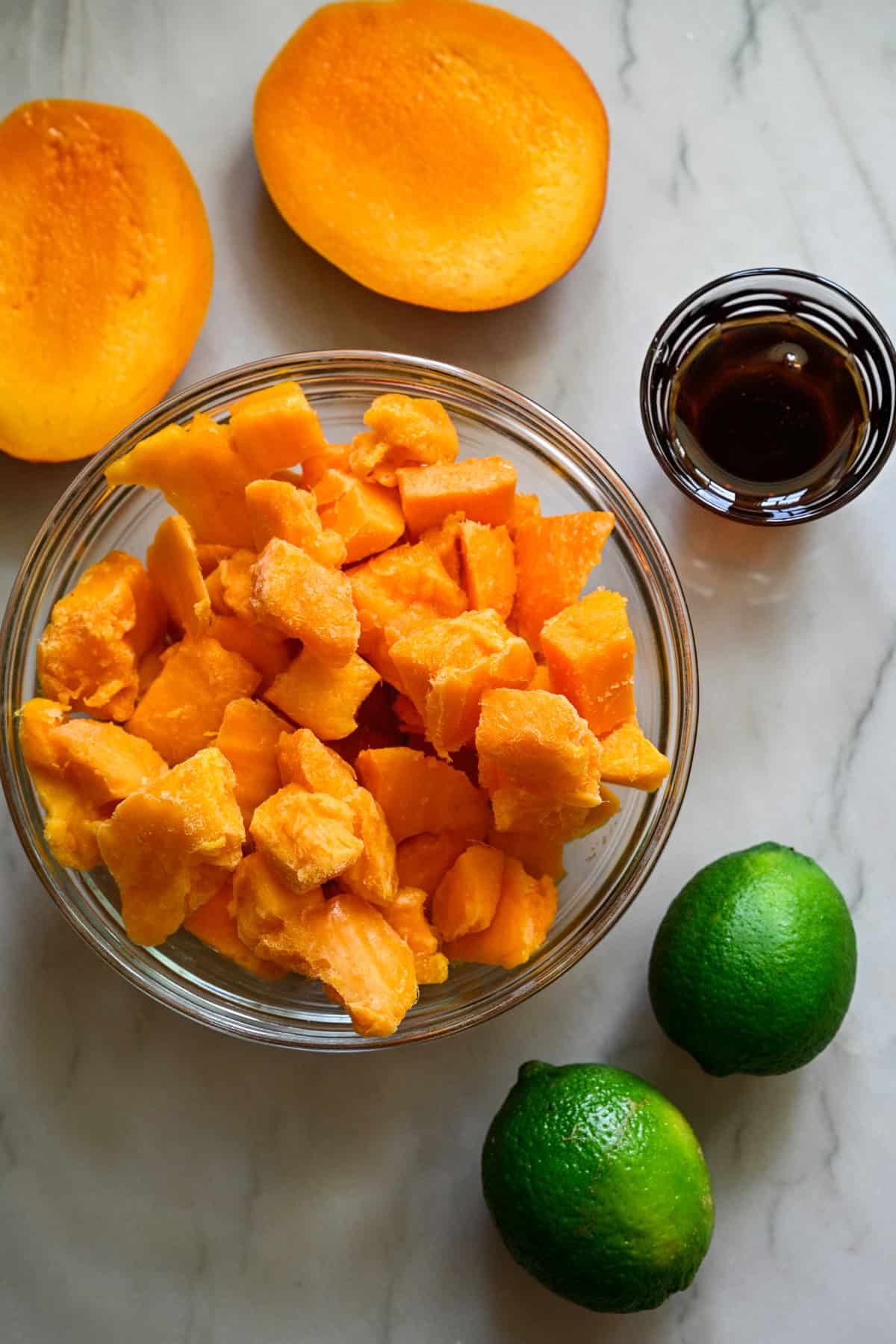 bovenaanzicht van in blokjes gesneden mango in een kom, 2 limoenen en agavesiroop op het aanrecht.