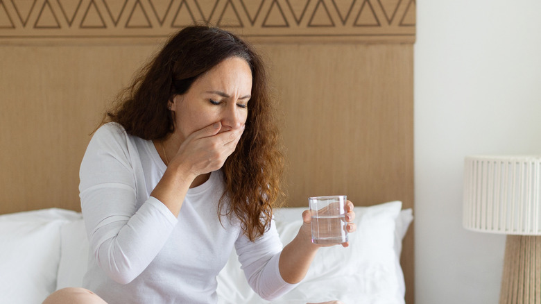 Een vrouw die zich misselijk voelt terwijl ze een glas water vasthoudt en op haar bed zit