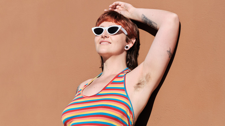 Een jonge vrouw die lacht en tegen een muur leunt terwijl ze in een tanktop met haar ongeschoren oksels pronkt