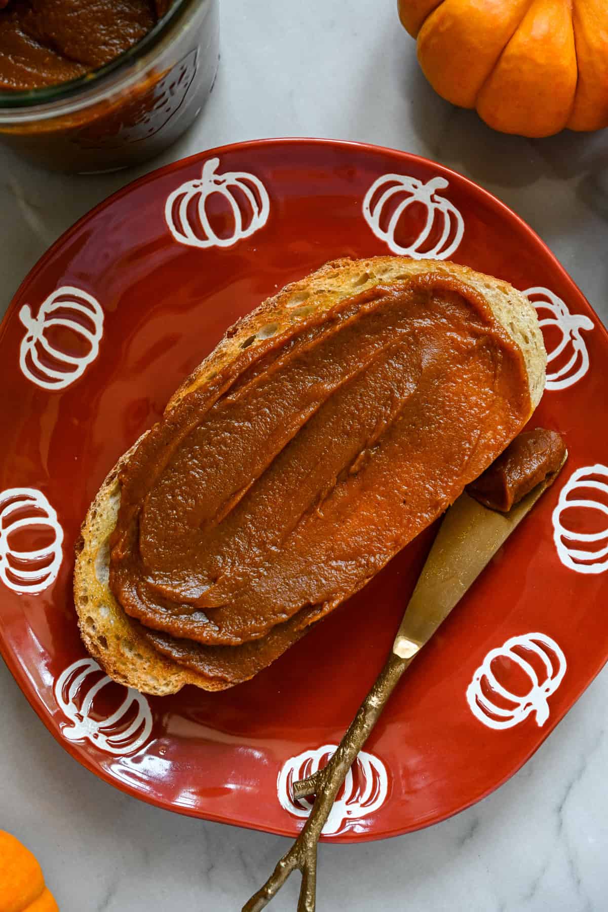 Een sneetje toast ingesmeerd met zelfgemaakte pompoenboter op een oranje bord met pompoenen langs de rand.
