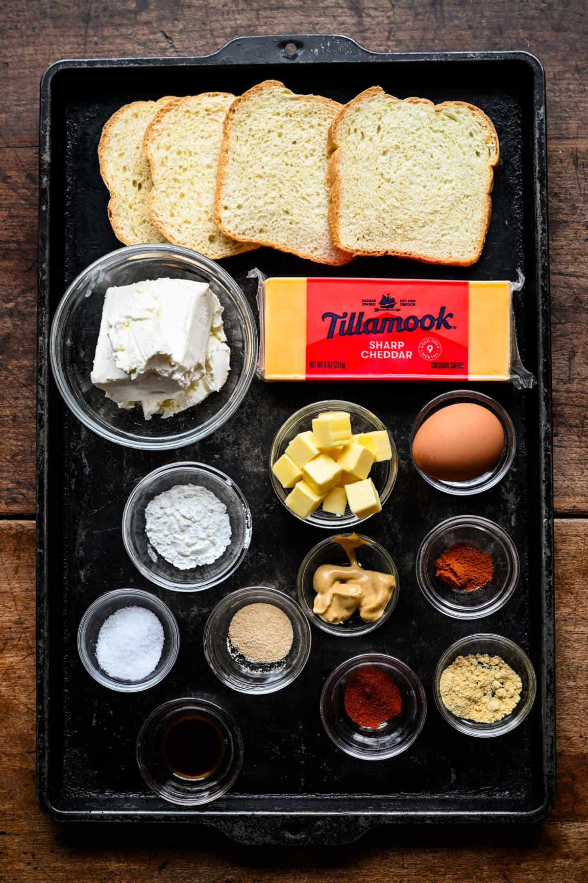 Bovenaanzicht van ingrediënten voor kaasdromen op een donkere bakvorm: brood, roomkaas, een blokje Tillamook scherpe cheddar, boterblokjes, een ei, mosterd en kleine kommen met kruiden.