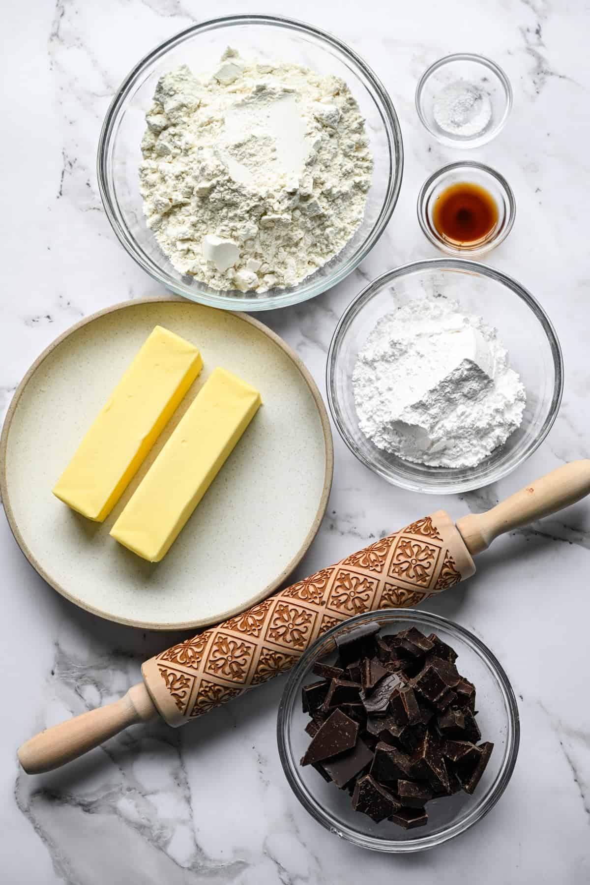 Glazen kommen met bloem, poedersuiker, gehakte pure chocolade, vanille en zout op een marmeren aanrecht met twee klontjes boter op een bord en een houten deegroller met reliëf.