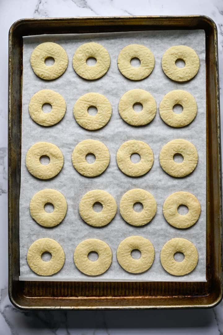 Twintig gebakken ringvormige zandkoekkoekjes, lichtgouden met een reliëfpatroon, op een met bakpapier beklede bakplaat.