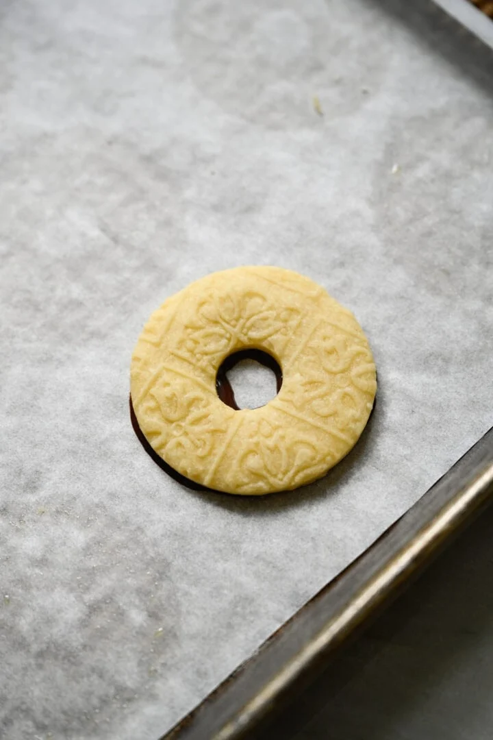Ringvormig zandkoekkoekje met reliëf, geperst op een ring van gesmolten chocolade op een met bakpapier beklede bakplaat.