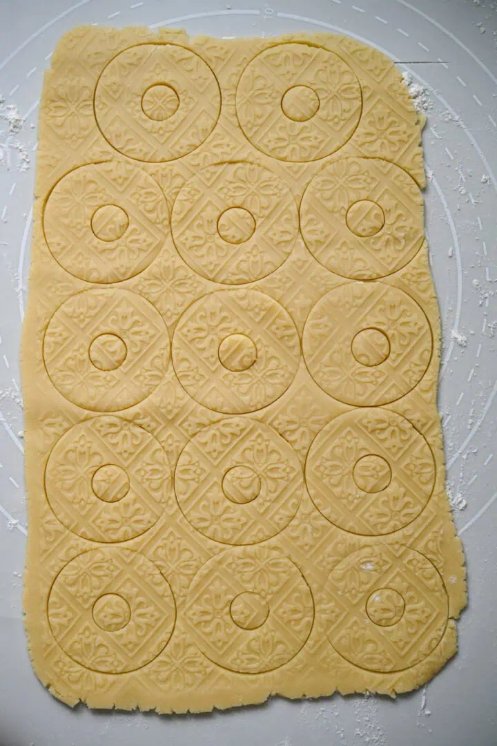 Uitgerold zandkoekdeeg met een bloementegelpatroon in reliëf en in rondjes met kleine gaatjes in het midden gesneden, voordat de koekjes eruit worden gehaald.
