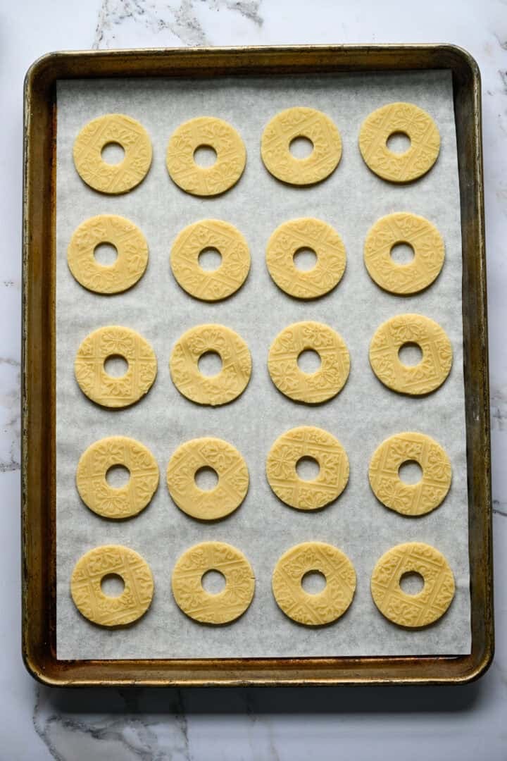 Twintig ongebakken ringvormige zandkoekjes met een reliëfpatroon, in rijen op een met bakpapier beklede bakplaat.