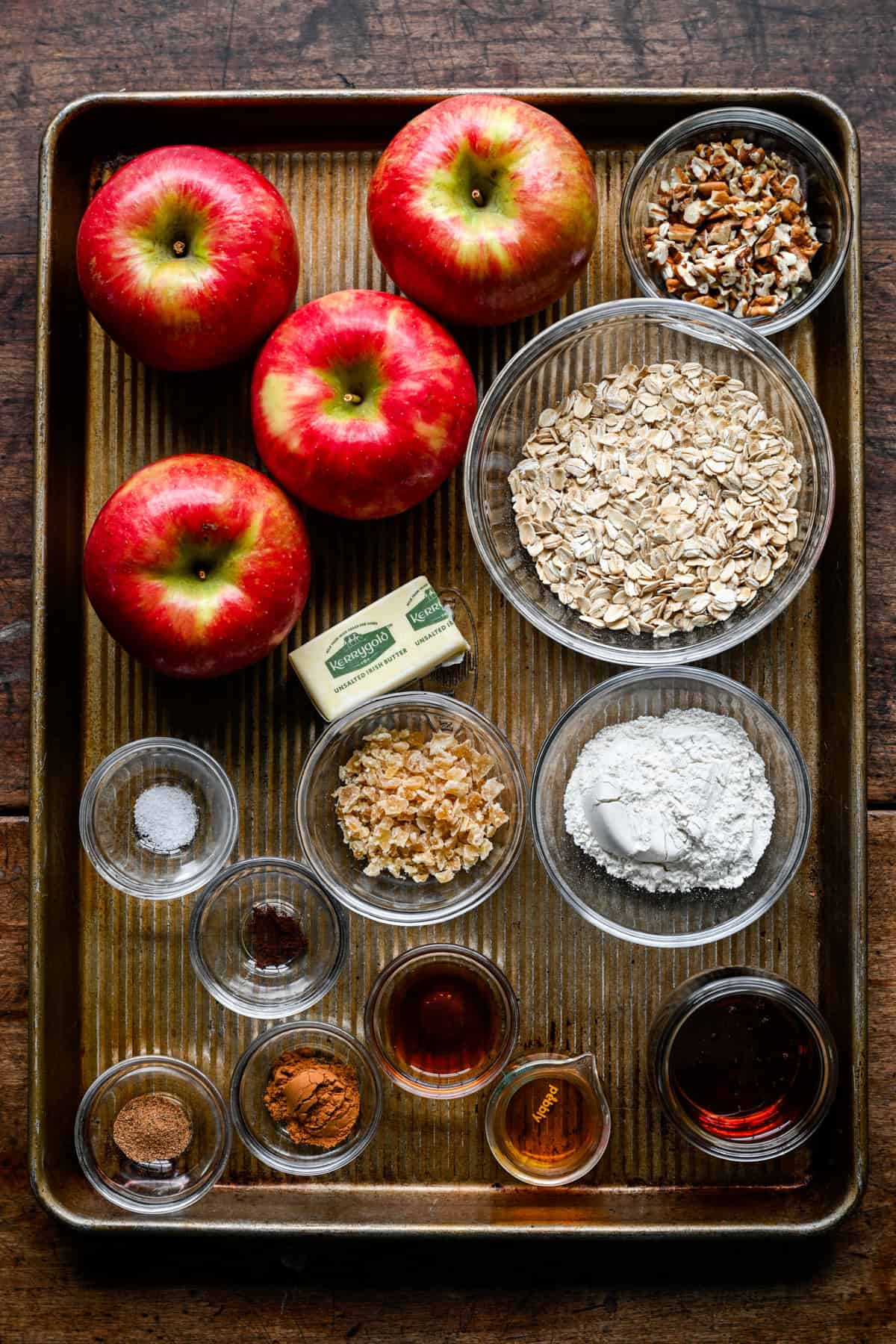 Ingrediënten voor gebakken appels op een bakplaat: vier honingkrokante appels, kommen havermout, gehakte pecannoten, bloem, gekristalliseerde gember, een stuk boter, gemalen kruiden, ahornsiroop en vanille.