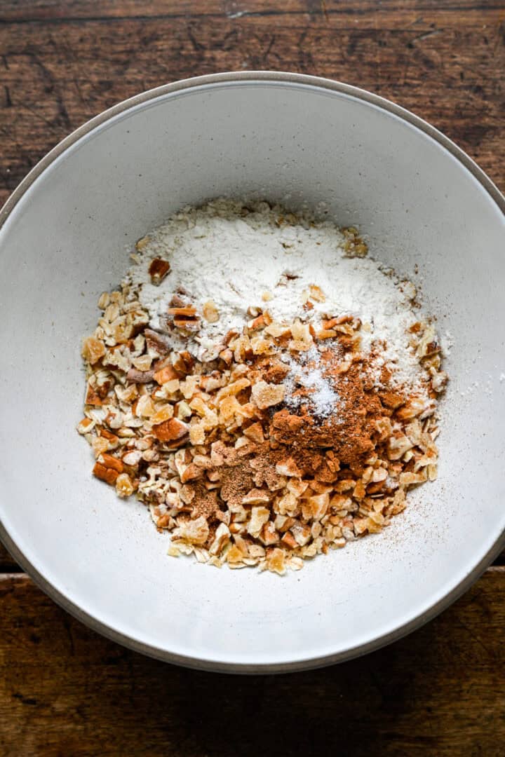 Gerolde haver, gehakte pecannoten, gekristalliseerde gember, bloem, kaneel, nootmuskaat, kruidnagel en zout in een witte mengkom voordat je roert.