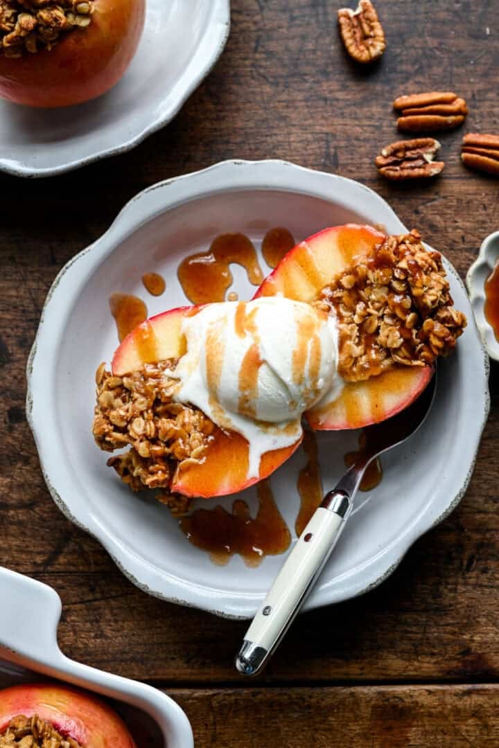 Een gehalveerde gebakken appel met vanille-ijs en dadelkaramel, een tweede portie en verspreide pecannoten op de achtergrond.