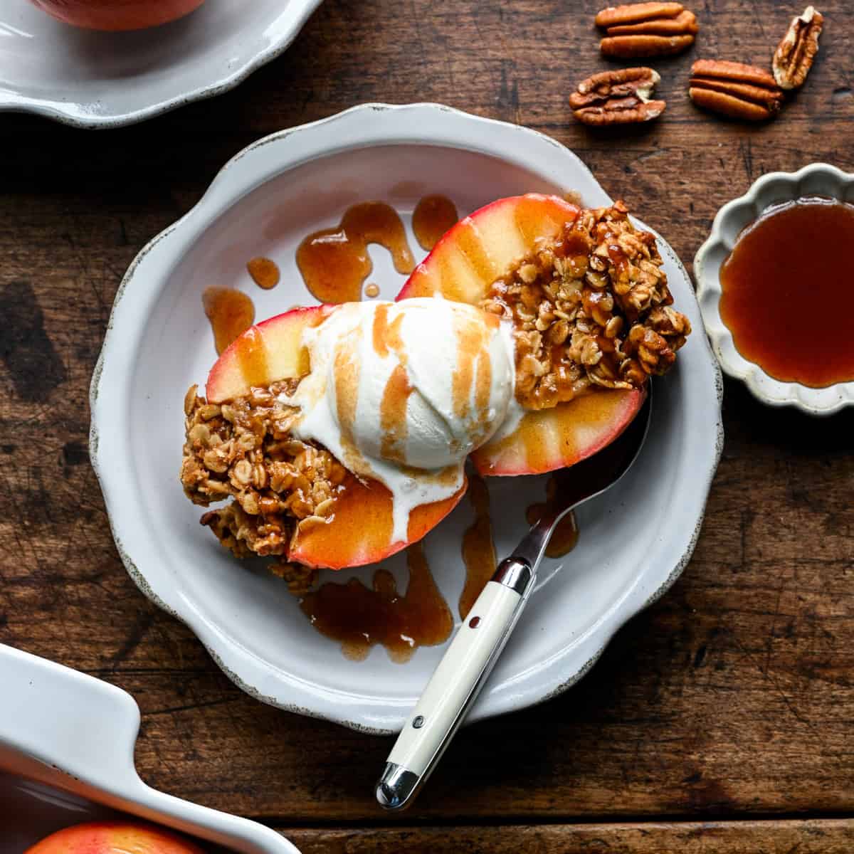 Een gebakken appel in tweeën gesneden op een bord met een bolletje vanille-ijs, besprenkeld met dadelkaramel, pecannoten en een klein schaaltje karamel ernaast.
