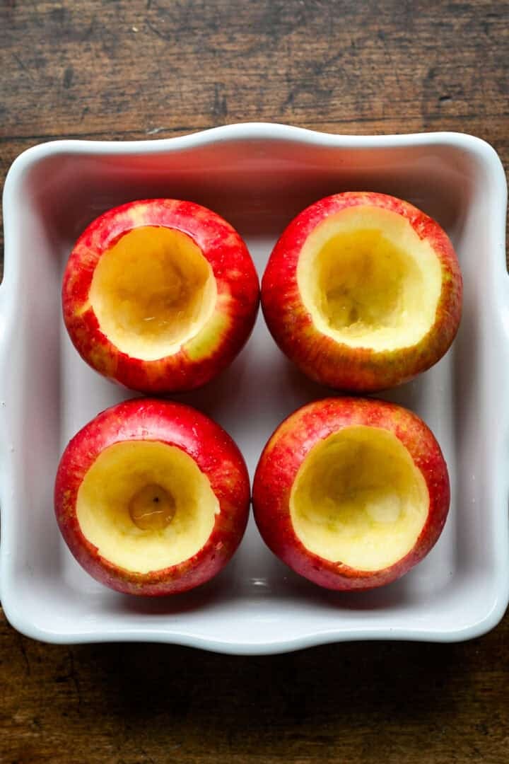 Vier honingkrokante appels, zonder klokhuis en met intacte bodem, staande in een witte vierkante ovenschaal.