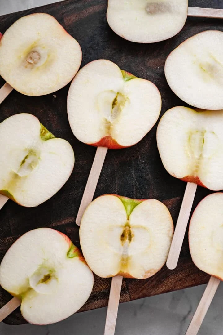 Bovenaanzicht van dikke appelschijfjes op ijslollystokjes op een donkere houten snijplank.