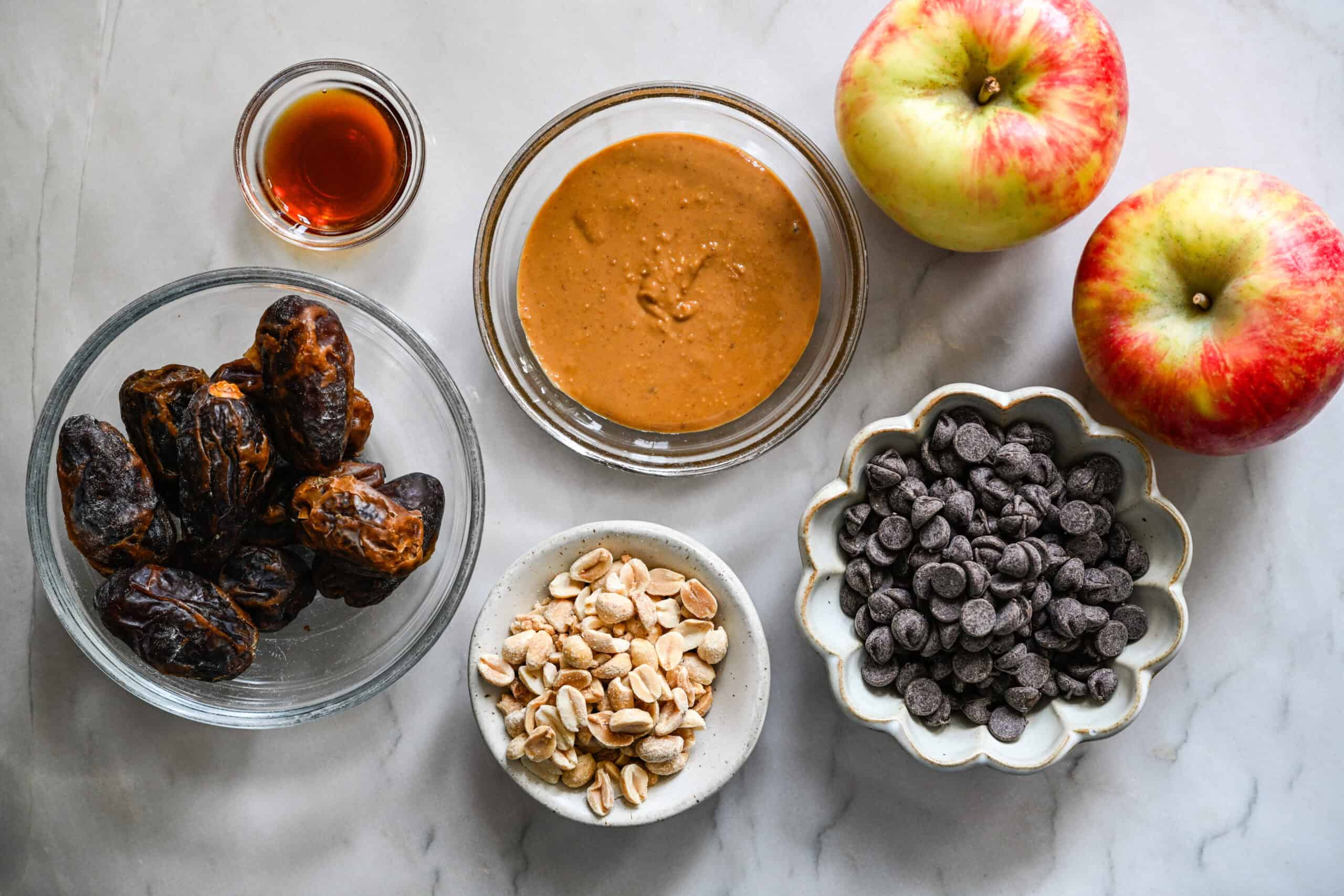 Bovenaanzicht van dadels, pindakaas, vanille, pinda's, chocoladestukjes en twee appels op een marmeren aanrecht.