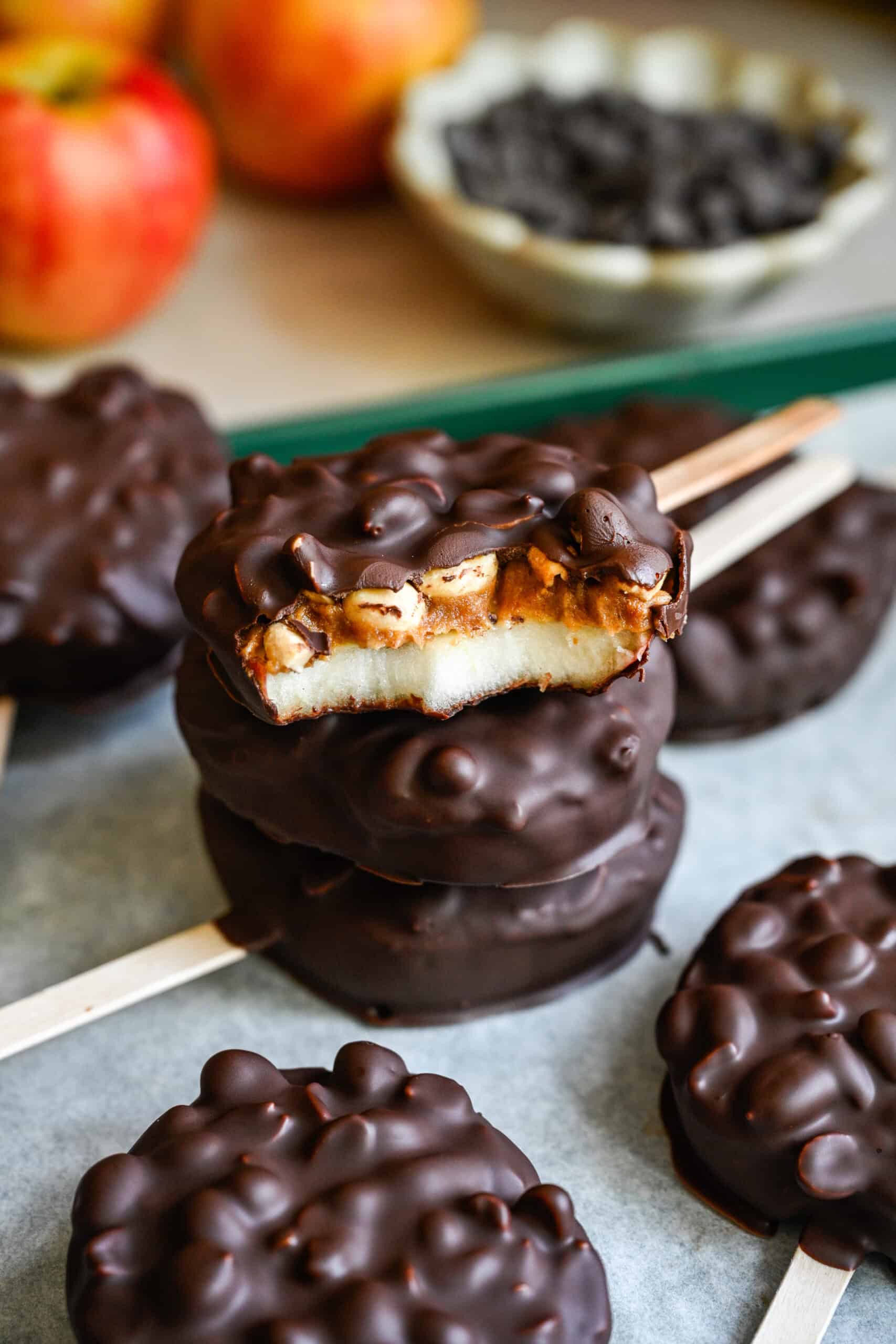 45 graden zicht op een stapel met chocolade bedekte appelschijfjes, waarvan de bovenste is gebeten om de appel, dadelpasta en pinda's te laten zien.