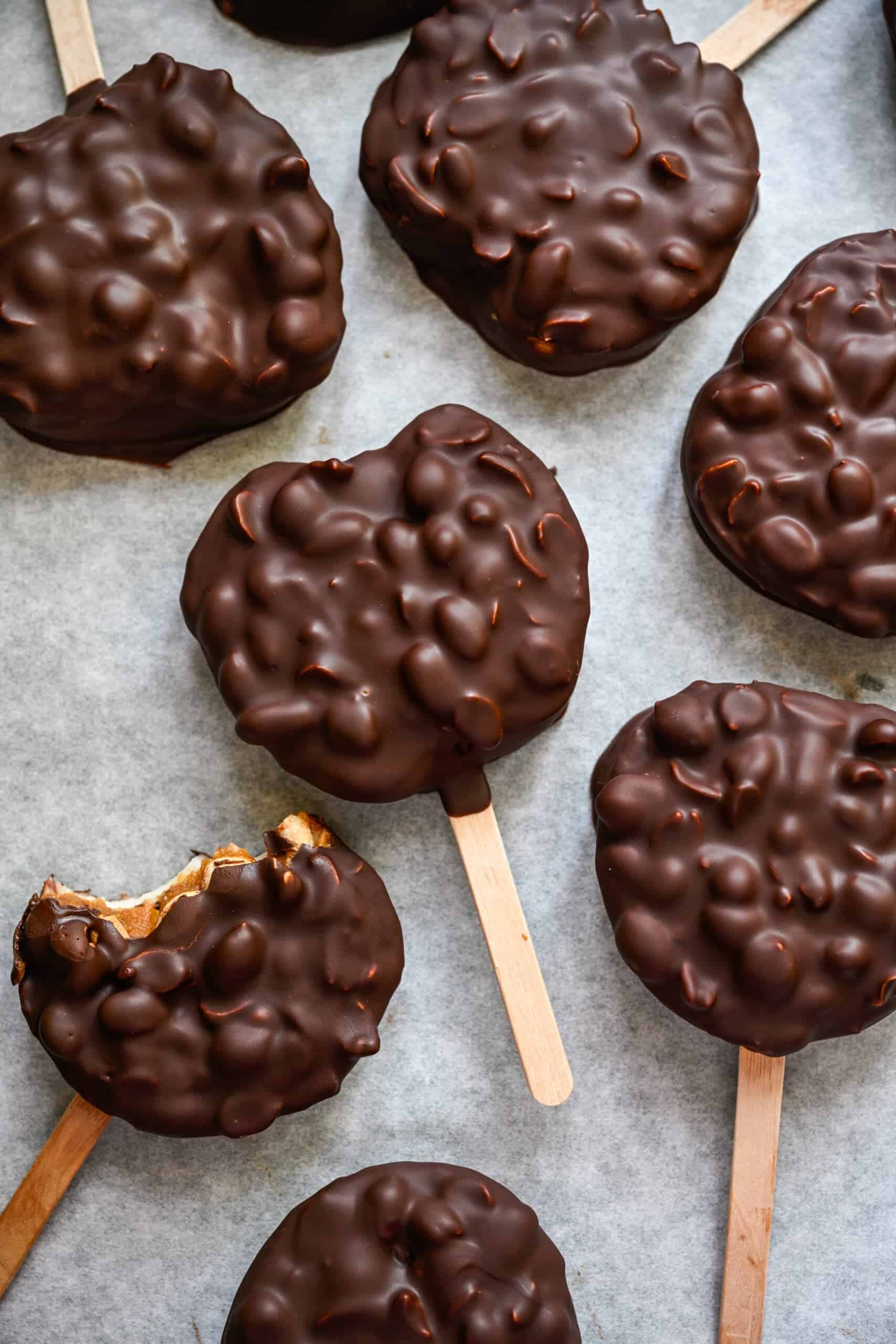 Bovenaanzicht van met chocolade bedekte appelschijfjes op ijslollystokjes op perkamentpapier, één met een hap eruit.