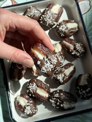 Close-up van een hand die een met chocolade bedekte dadel gevuld met kokosvulling uit een dienblad tilt.