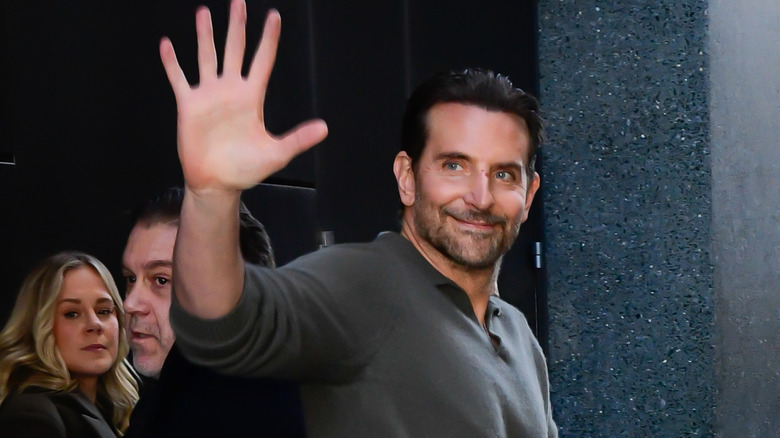 Bradley Cooper zwaait en glimlacht terwijl hij door een drukke straat loopt