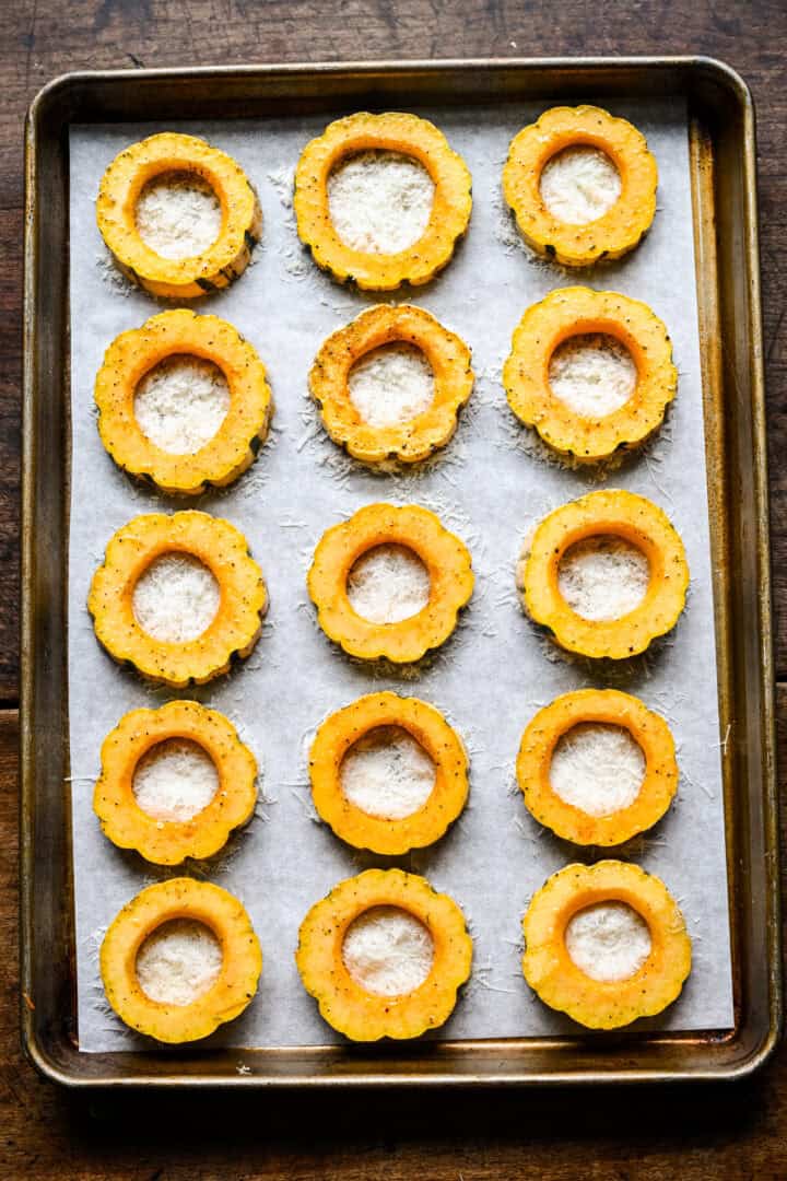 Bovenaanzicht van gekruide delicata-pompoenringen op een met perkament beklede bakvorm, elk gevuld met geraspte Parmezaanse kaas.