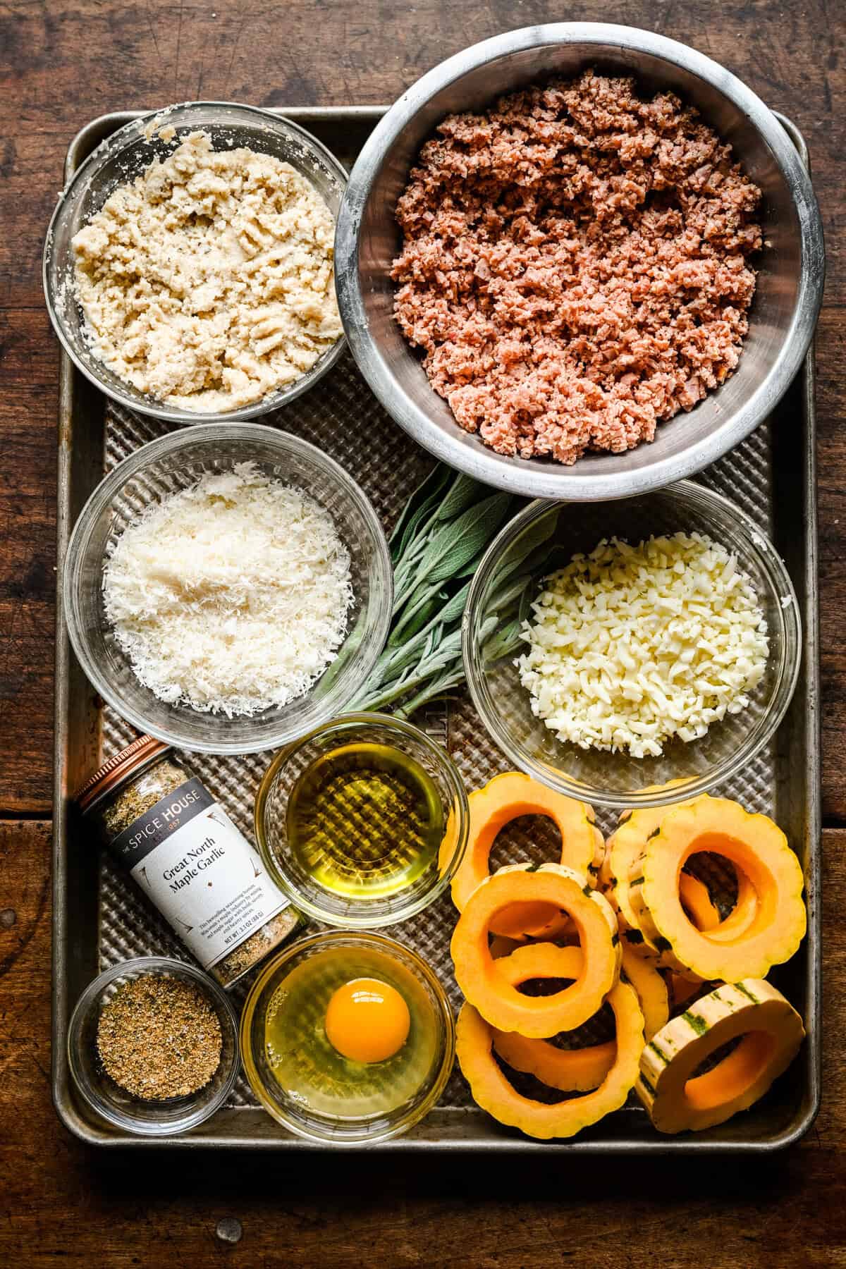 Bovenaanzicht van ingrediënten op een bakplaat: gemalen worst, paneermeel, parmezaanse kaas, mozzarella, verse salie, olijfolie, een ei, Great North Maple Garlic-kruiden en delicata-pompoenringen.