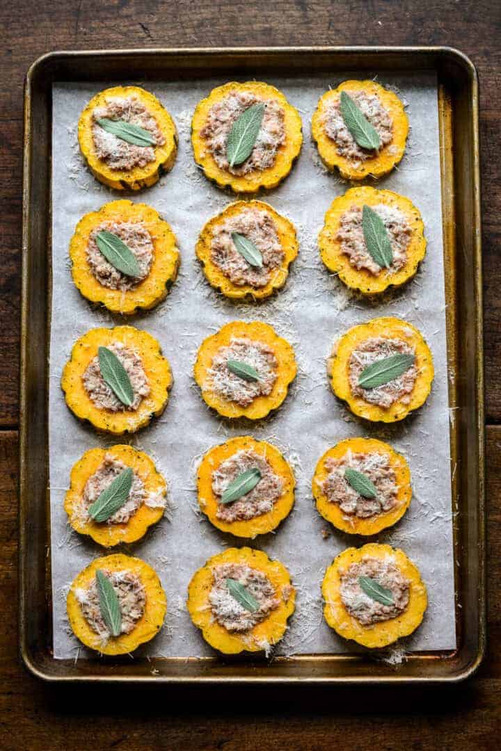 Bovenaanzicht van squashringen op een bakplaat gevuld met worstvulling, bestrooid met Parmezaanse kaas en gegarneerd met salieblaadjes voor het bakken.