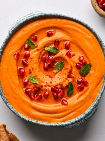 Bovenaanzicht van wortel en geroosterde feta-dip van rode paprika in een blauwe kom, gegarneerd met granaatappelpitjes en munt, met pitabroodje en kleine kommen granaatappelpitjes en munt aan de zijkant.