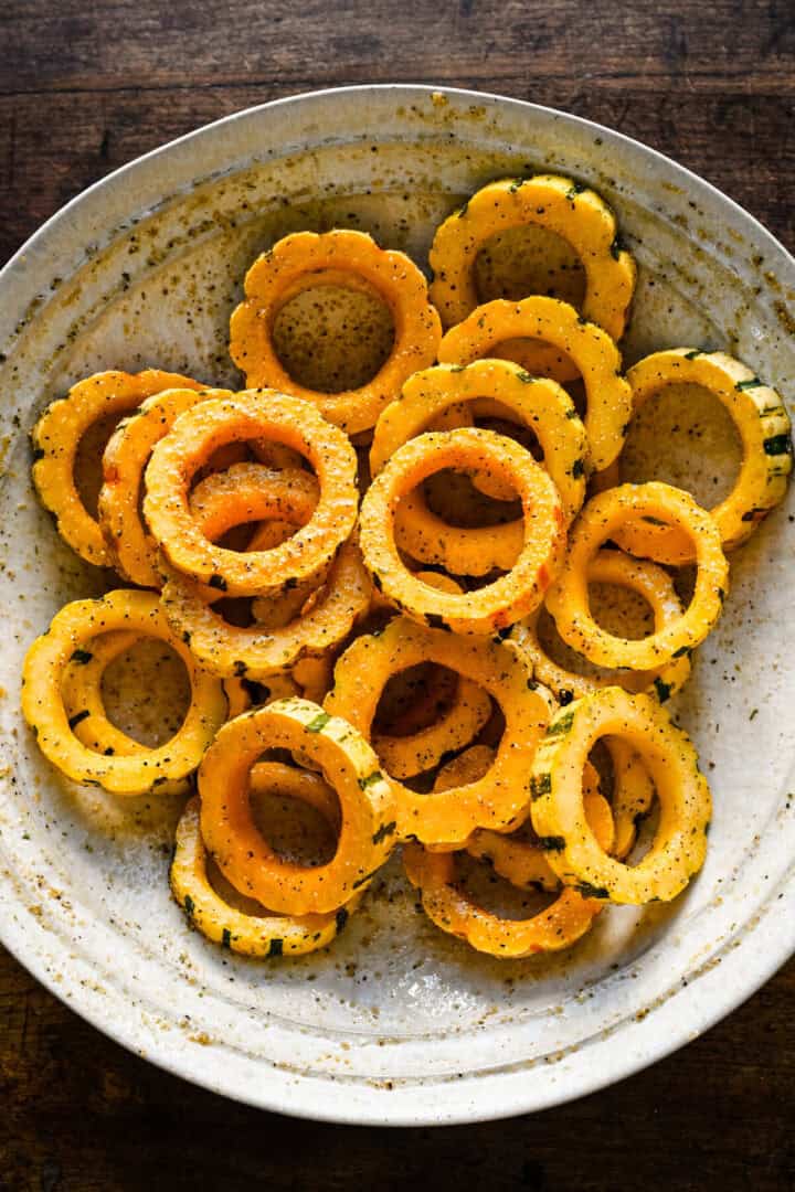 Bovenaanzicht van delicata squashringen gegooid met olie en kruiden in een witte kom.