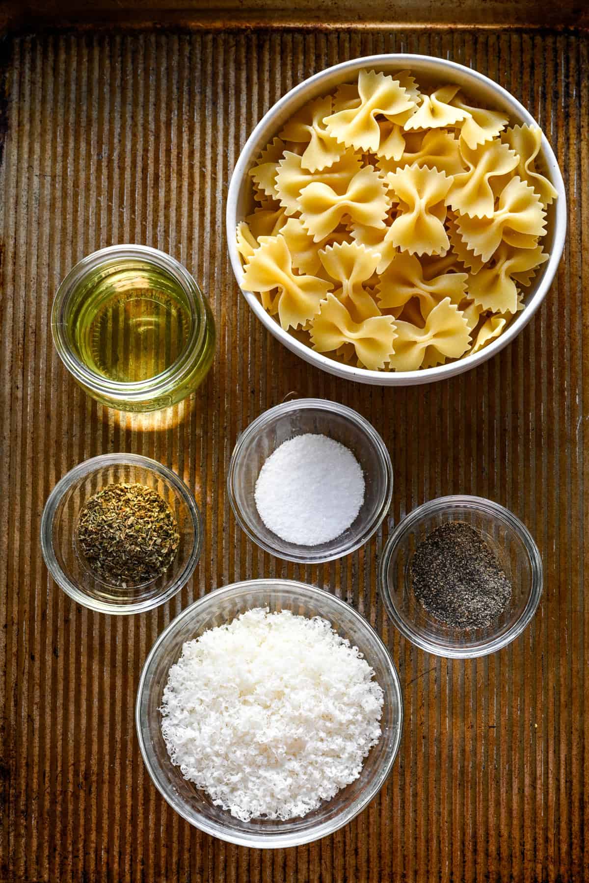 Bovenaanzicht van ingrediënten voor pastabladerdeeg op een bladpan: vlinderdaspasta, olie, gedroogde kruiden, zout, peper en geraspte Parmezaanse kaas.