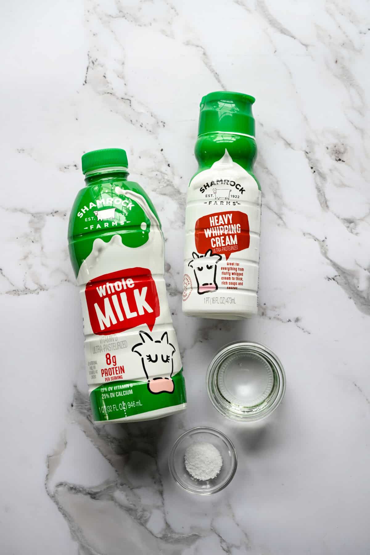 Bovenaanzicht van ingrediënten voor zelfgemaakte kwark: een fles Shamrock Farms volle melk, Shamrock Farms zware slagroom en kleine glazen kommen witte azijn en zout op marmer