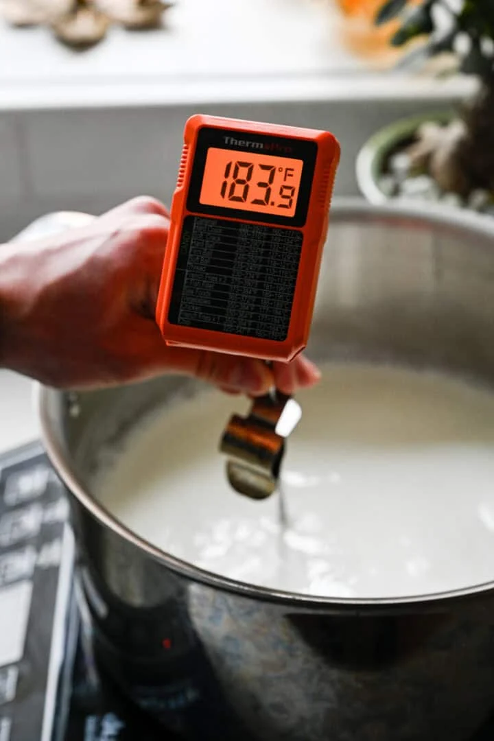 Hand die een ThermoPro-thermometer vasthoudt en 183 ° F aangeeft boven een pan met verwarmende melk