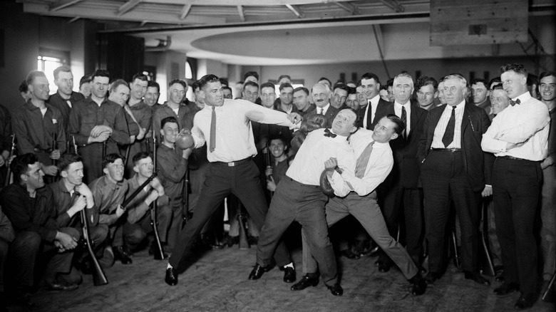 Harry Houdini zet zich schrap voor impact terwijl bokser Benny Leonard en Jack Dempsey zich voorbereiden op een swing