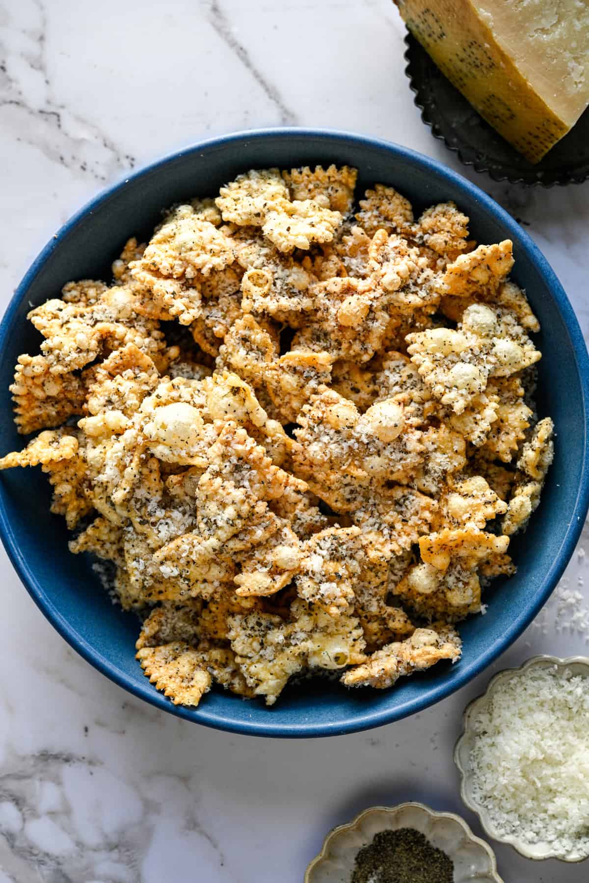 Bovenaanzicht van gepofte pasta in een blauwe kom naast een stukje Parmezaanse kaas en een schaaltje peper.